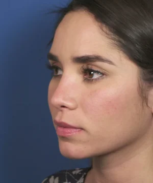 A woman with dark hair and a serious expression looks to the side against a blue background.