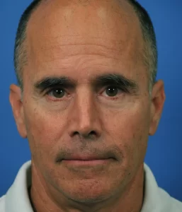 A middle-aged man with a neutral expression is shown against a blue background, wearing a white collared shirt.