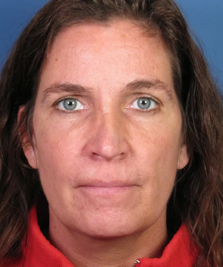 A woman with long brown hair and a neutral expression, wearing a red top, against a blue background.