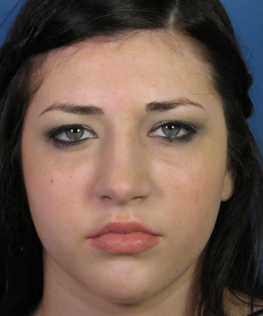 A person with long dark hair and green eyes against a blue background, looking directly at the camera with a neutral expression.