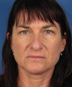 A middle-aged person with straight brown hair and a serious expression against a blue background.