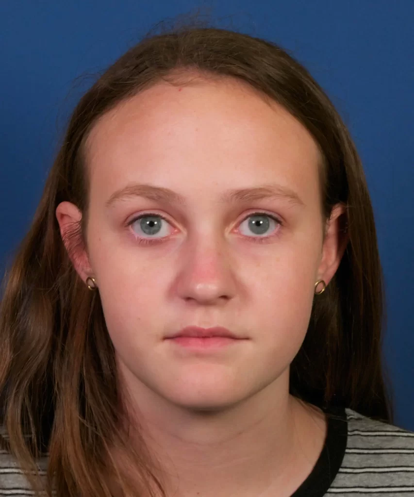 A person with long brown hair and blue eyes is looking directly at the camera against a blue background, wearing a striped top and small hoop earrings.