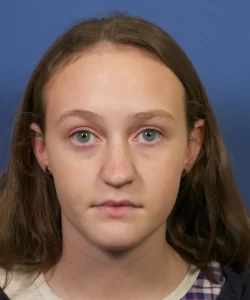 A person with long brown hair and blue eyes looks directly at the camera against a blue background.