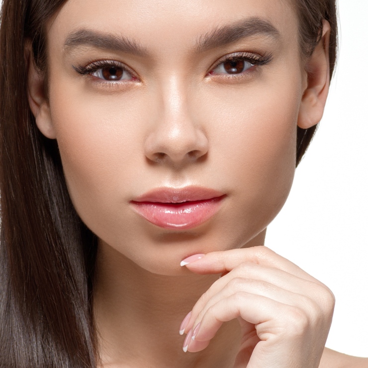 Close-up of a woman with long brown hair, touching her chin, and looking directly at the camera. She has clear skin and is wearing minimal makeup.