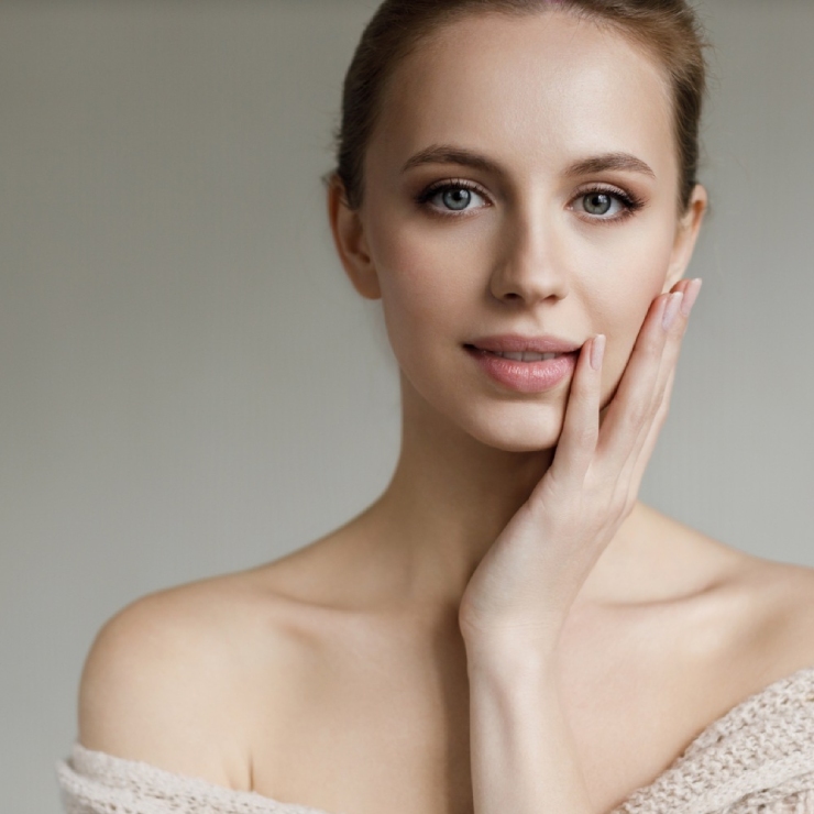 Woman with light hair and bare shoulders touching her face, looking at the camera against a neutral background.