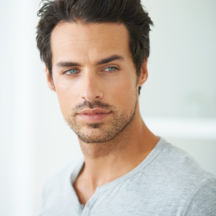 Man with dark hair and stubble wearing a light gray shirt, looking slightly to the side against a soft, light background.