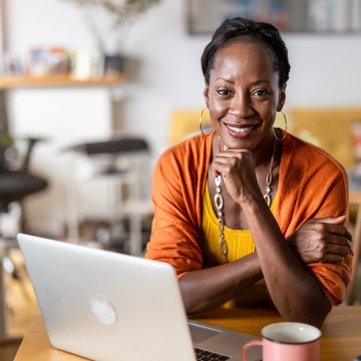 A person sitting at a desk with a laptop and a mug, smiling, wearing a yellow shirt and orange cardigan.