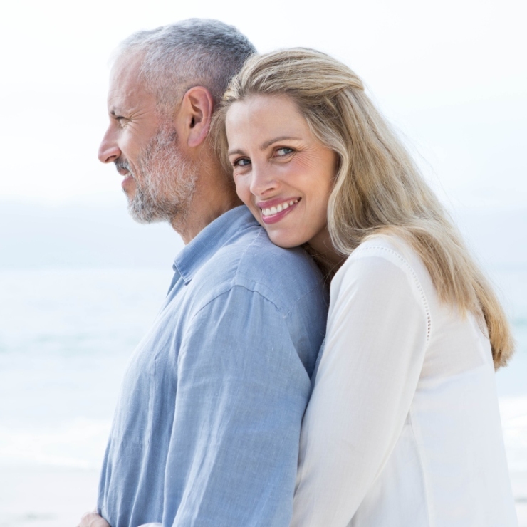 A woman embraces a smiling man from behind at a beach.