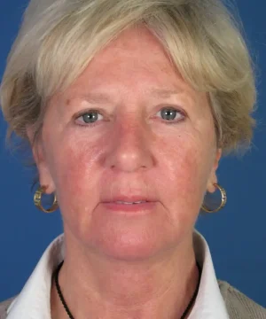 Portrait of a person with short blonde hair against a blue background, wearing a white collared shirt, a black necklace, and gold hoop earrings.