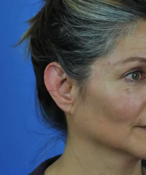 Side profile of a woman with dark and gray hair pulled back, against a blue background.