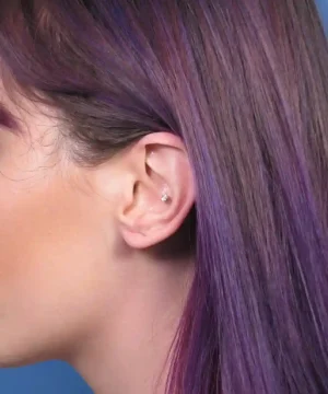 Close-up of a person's ear with a small stud earring, hair dyed purple, against a blue background.