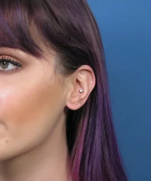 Person with purple hair showing a helix ear piercing against a blue background.