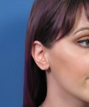 Close-up of a woman's side profile with purple hair, focusing on her ear and eye against a blue background.