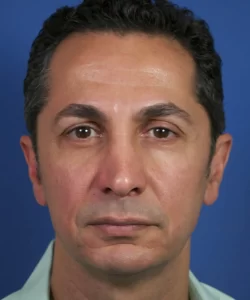 A man with short, curly dark hair and wearing a light green shirt against a blue background.