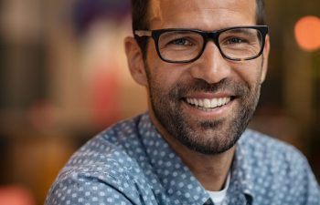 Man with glasses and a beard wearing a patterned shirt, smiling in a blurred indoor setting.