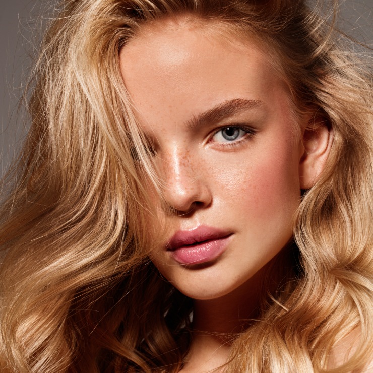 A person with long blonde hair and blue eyes looks directly at the camera. They have a neutral expression and subtle makeup.