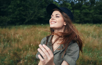 Woman in a black cap and green shirt smiling in a grassy field with trees in the background.