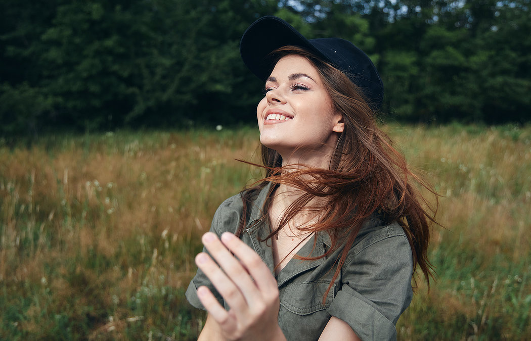 Woman in a black cap and green shirt smiling in a grassy field with trees in the background.