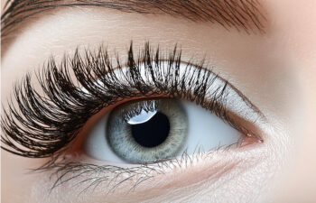 Close-up of an eye with long eyelashes, greyish-blue iris, and well-defined eyebrow above, showcasing detailed eye makeup.