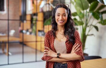 A person with long hair, wearing a patterned shirt and glasses on their head, smiles confidently indoors with houseplants in the background.