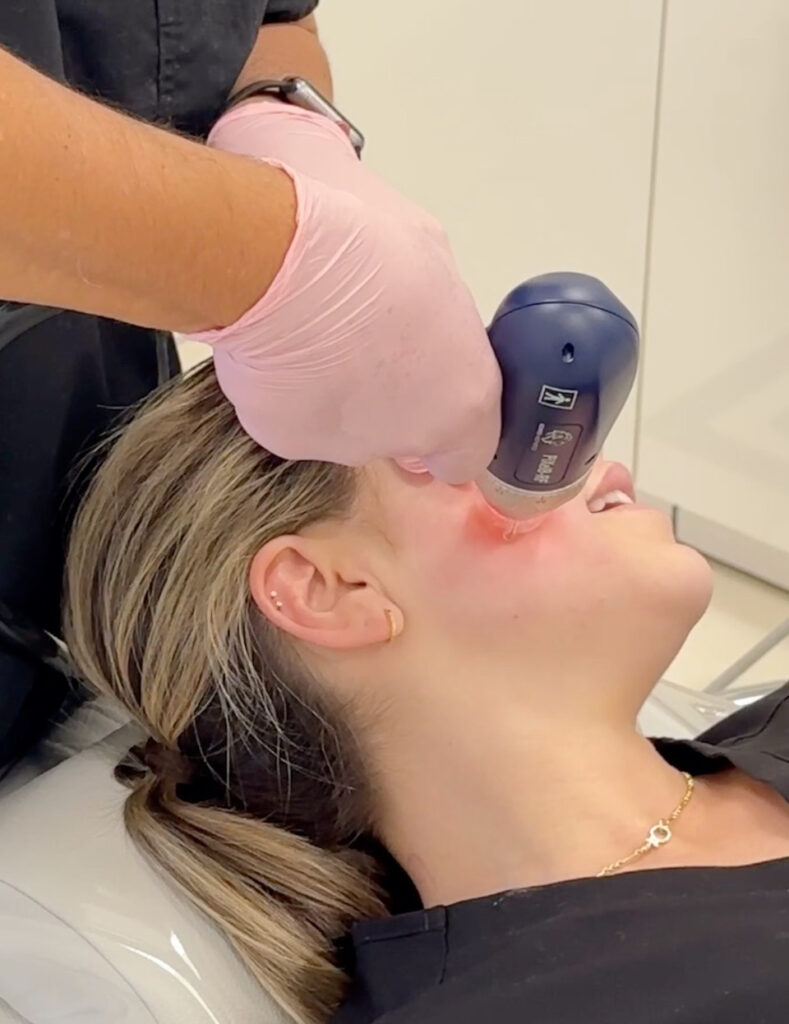 A person receives a facial laser treatment from a professional wearing pink gloves, targeting the area around the cheekbone and eye.