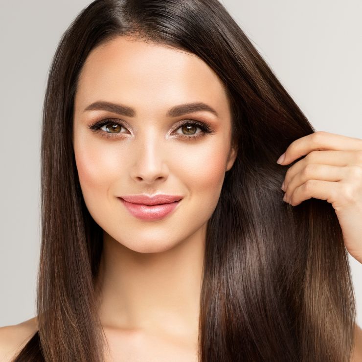 A woman with long, straight brown hair and natural makeup smiles slightly while touching her hair against a simple, light background.