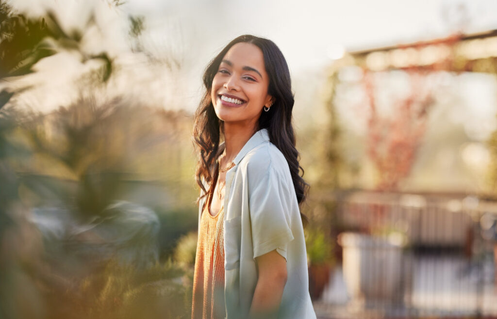 Woman with long dark hair smiling outdoors, standing surrounded by greenery and blurred plants, wearing a light shirt over an orange top on a sunny day.